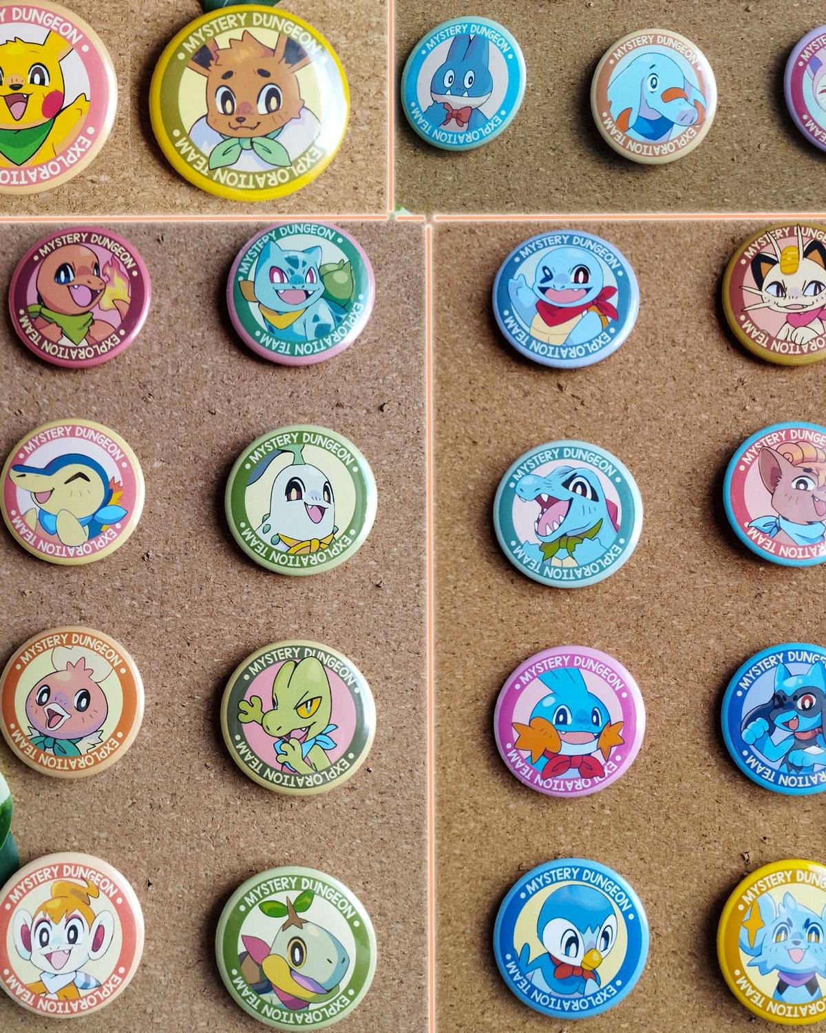 Une vingtaine de badges ronds épinglés sur des tableaux en liège : chaque badge montre un Pokémon souriant avec un foulard, entouré d'un anneau coloré.