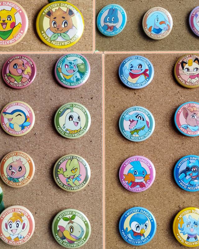 Une vingtaine de badges ronds épinglés sur des tableaux en liège : chaque badge montre un Pokémon souriant avec un foulard, entouré d'un anneau coloré.